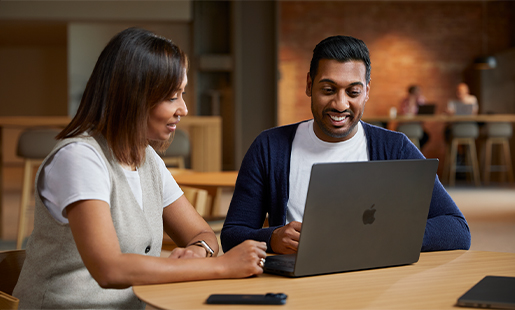 Amar colaborando con una compañera, sentado con su MacBook en una mesa de una zona común.