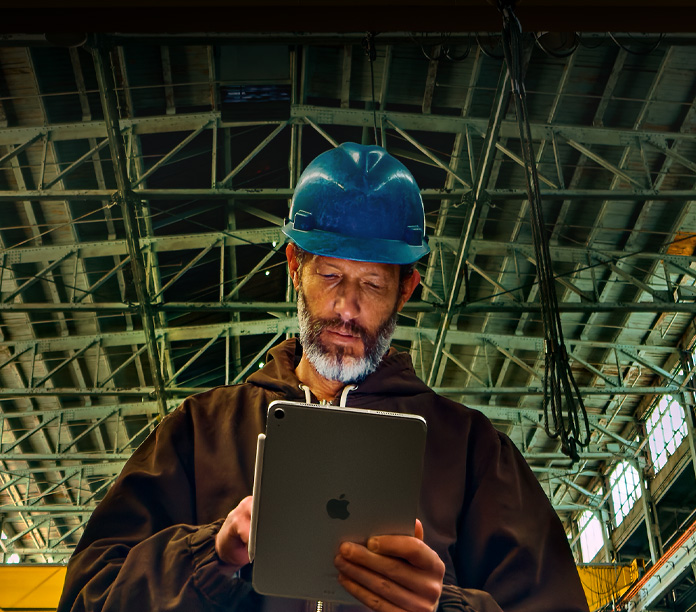 一個人身穿棕色外套、頭戴藍色安全帽，手持貼合著 Apple Pencil 的銀色 iPad，身處工業環境中工作，上方可見金屬橫樑