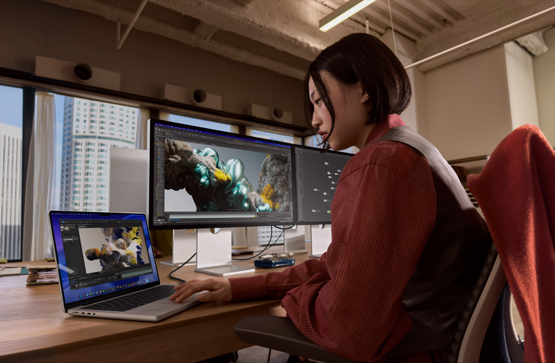 一個人正在辦公桌前工作，MacBook Pro 連接到外部顯示器，兩個螢幕均顯示 3D 算圖，背景的窗外可見建築物