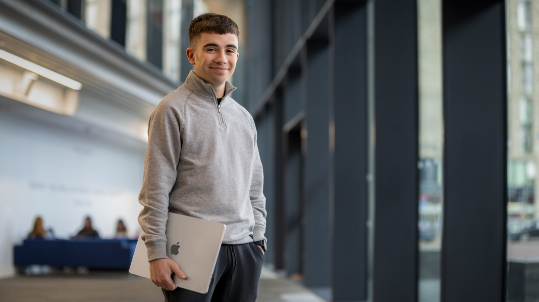 Un étudiant, debout dans le couloir d’un bâtiment moderne, tient un ordinateur portable MacBook, en souriant à l’objectif