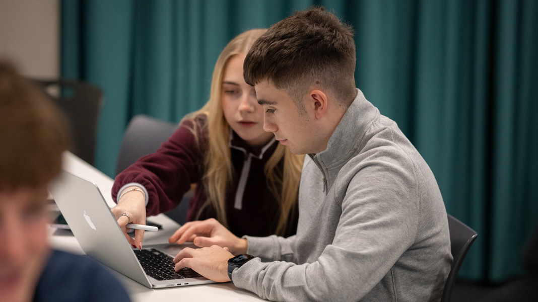 Deux camarades de classe travaillent ensemble sur un MacBook, elle pointe l’écran du doigt pendant qu’il tape