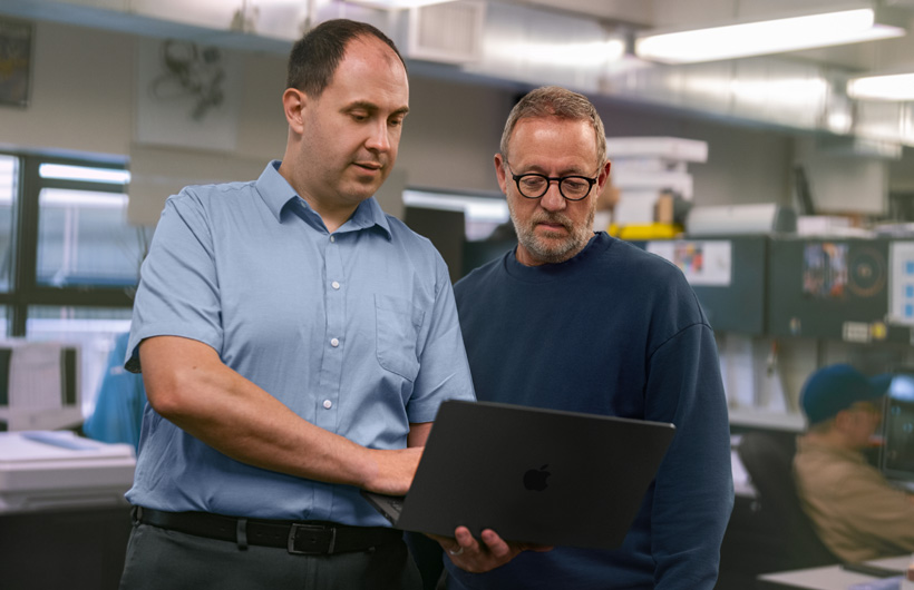 Dois homens em uma estação de trabalho olham para um MacBook juntos. Um segura o aparelho, e o outro aponta para a tela