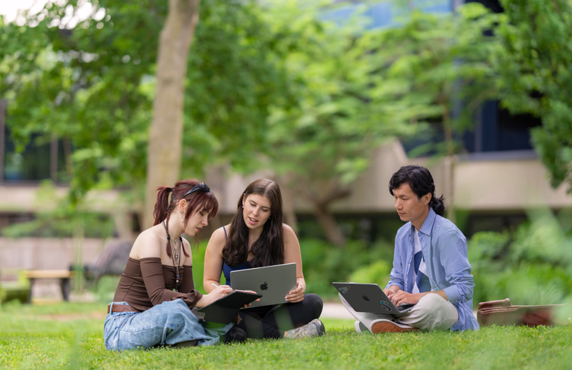 Três estudantes sentam-se na grama em um campus ao ar livre. Uma usa um MacBook, e a outra observa