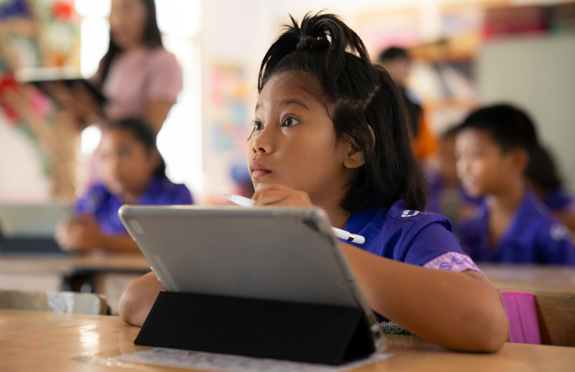 Une petite fille est assise à son bureau, elle regarde vers l’avant de la classe avec beaucoup d’attention. Elle utilise un iPad et tient un Apple Pencil.