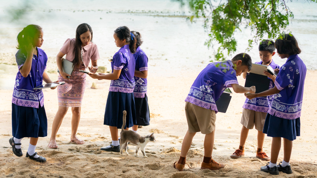 Six élèves de l’école primaire portant des uniformes violets colorés sont debout sur une plage de sable et tiennent des iPad. Une enseignante aide une élève sur un iPad, et un chat déambule au milieu.