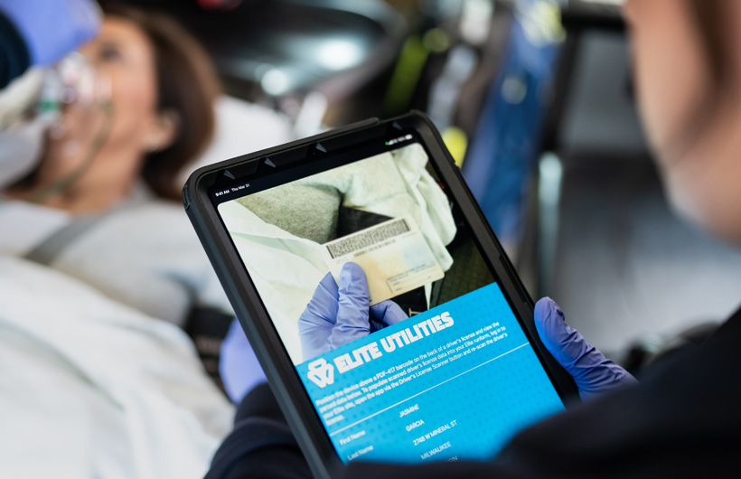 Elite Utilities-appen er åben på iPad, mens en paramediciner scanner en patients kørekort, og patienten ligger på en båre
